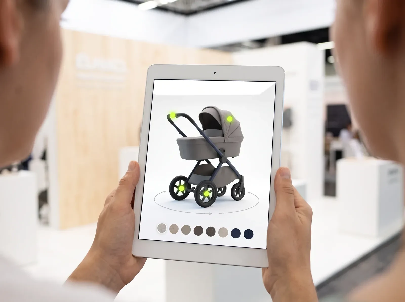 Hands hold a tablet at a trade-fair booth; the screen shows a 3D stroller with glowing hotspots and a row of fabric swatches